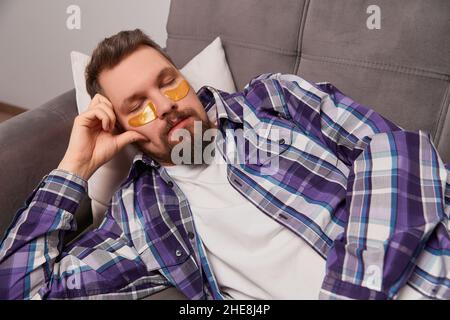 Homme dans une chemise violette avec des yeux patchs dormant sur le canapé.Auto-soin de routine Banque D'Images