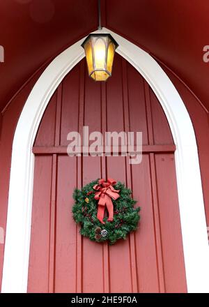 La couronne de Noël festive sur une porte d'église en bois rouge accueille la saison de l'Avent.Une lumière dorée surplombe la porte. Banque D'Images