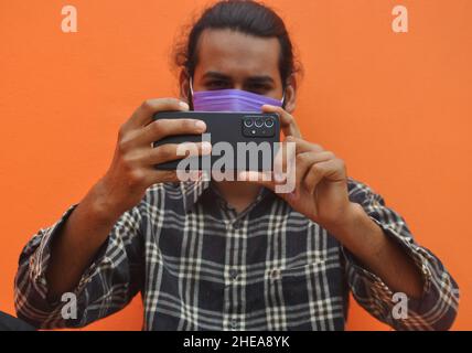 Mise au point sélective d'un jeune homme portant un masque facial en prenant des photos de vous avec son téléphone mobile, posant contre un mur de couleur orange avec un espace de copie Banque D'Images