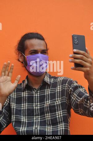 Gros plan d'un jeune homme portant un masque, en lui disant bonjour en lui faisant passer la main pendant un appel vidéo, en se posant contre un mur orange. Banque D'Images
