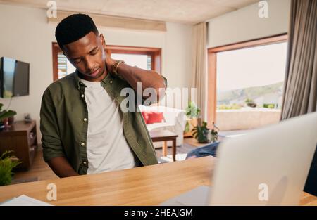 Jeune homme noir adulte tenant son cou dans la douleur pendant qu'il travaille sur ordinateur portable de la maison dans la cuisine moderne. Banque D'Images