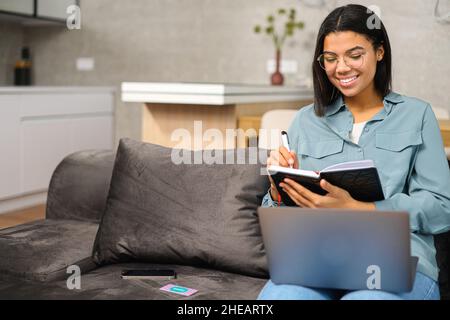 Une jeune fille adolescente en verre souriante, assise sur le canapé avec un ordinateur portable sur les genoux et prenant des notes tout en étudiant à la maison Banque D'Images