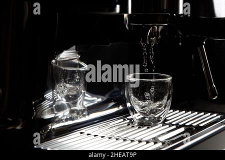 Gros plan de gouttes d'eau chaude qui s'écoulant dans une tasse à espresso transparente provenant d'une machine à café manuelle pour le préchauffage, monochrome, avec espace de copie Banque D'Images