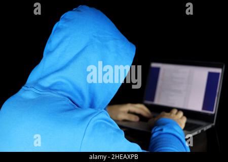 Cybercriminalité, piratage et criminalité technologique.Homme à capuche bleu assis avec un ordinateur portable sur fond noir, concept de piratage de torrent et contenu illégal Banque D'Images