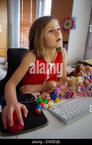 Une fille de six ans pratiquant la lecture sur un ordinateur à la maison, accompagnée de nombreuses poupées Banque D'Images