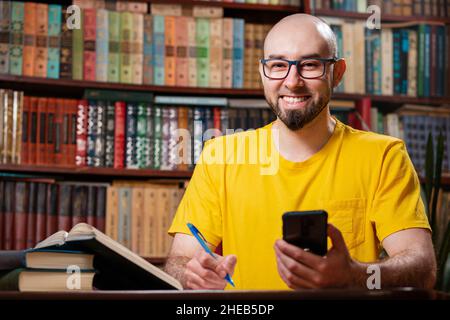 Un homme à barbe avec des lunettes utilise un smartphone et écrit quelque chose dans un ordinateur portable.Étagères en arrière-plan.Le concept d'un réseau social Banque D'Images