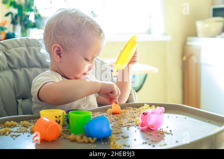 petit enfant assis sur une chaise d'alimentation et joue avec des ustensiles de jouet et du grain. développement de compétences en moteur fin chez les enfants jusqu'à l'année. Développement précoce Banque D'Images