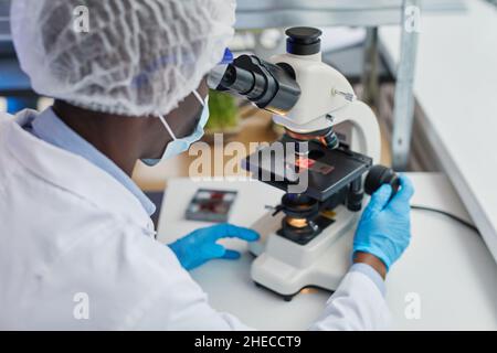 Vue arrière d'un médecin africain regardant à travers le microscope à la table, il examine un nouvel échantillon de virus Banque D'Images