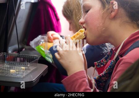 Enfant de neuf ans / enfant / fille mordu dans un fromage et un oignon pâteux qu'elle mange; pasties / remplir la nourriture avec beaucoup d'énergie pour les enfants, sur une table de plateau de siège, sur un Airbus a 320 ou un avion / avion / avion / avion 319.Une partie d'un panier repas pour elle préparé par un parent passager qui a pris une collation à bord du vol EasyJet pour une partie d'un repas indépendant.(128) Banque D'Images