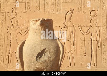 Égypte, haute Égypte, vallée du Nil, Edfu, statue de granit d'Horus devant l'entrée de la grande salle hypostyle du temple d'Horus avec en bas un bas-relief représentant sur la droite Hathor et Horus Banque D'Images