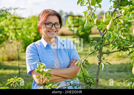 Portrait d'une femme d'âge moyen souriante et confiante avec bras croisés dans un verger, près d'un poirier Banque D'Images