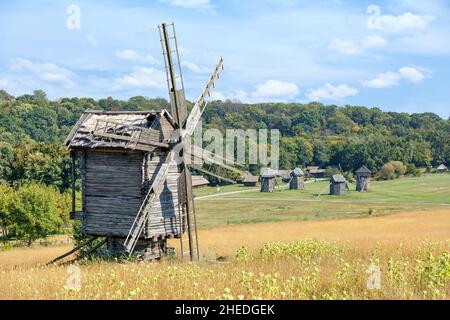 Un ancien moulin à vent en bois en bois abîmé se dresse sur la pente d'un champ rural surplombant un pré vert et une forêt dense. Banque D'Images