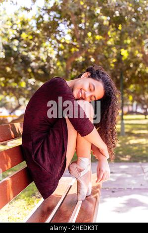 Jeune belle ballerine en robe noire assise sur un banc dans un parc avec ses yeux fermés et souriant Banque D'Images