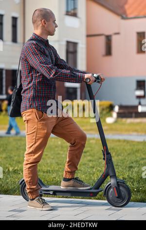 Jeune homme dans un pantalon et une chemise décontractés debout à côté de son scooter électrique Banque D'Images