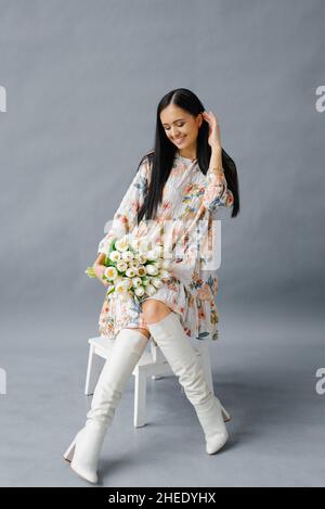 Une jeune jolie femme vêtue d'une robe et de bottes blanches est assise sur une chaise blanche avec un bouquet de tulipes blanches sur fond gris Banque D'Images