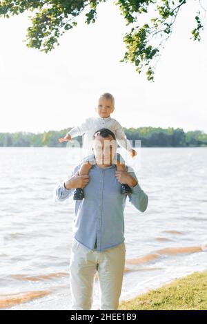 Le petit fils est assis sur le cou de papa.La famille marche au bord du lac en été Banque D'Images