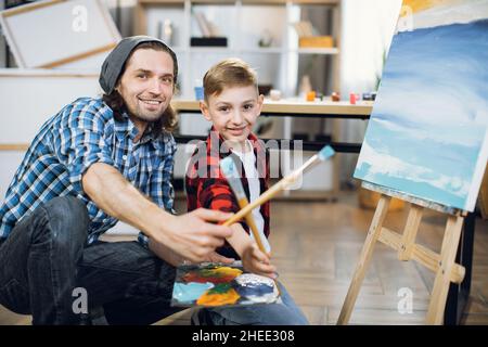 Beau caucasien homme et mignon garçon regardant la caméra tout en tenant dans les mains palette de couleurs et pinceaux.Portrait d'enseignant et d'étudiant à l'école d'art. Banque D'Images