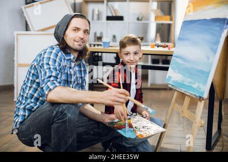 Beau caucasien homme et mignon garçon regardant la caméra tout en tenant dans les mains palette de couleurs et pinceaux.Portrait d'enseignant et d'étudiant à l'école d'art. Banque D'Images