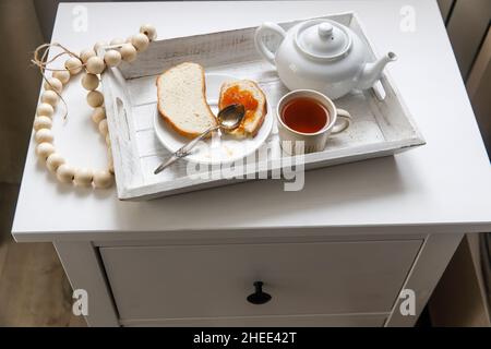 Une théière, une tasse de thé, deux tranches de pain grillé avec confiture d'abricot sur un plateau sur la table de chevet. Banque D'Images