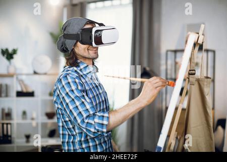 Un peintre caucasien portant un casque VR tout en peignant avec une brosse sur une toile en studio.Jeune homme utilisant une technologie innovante pour le processus créatif à l'intérieur. Banque D'Images