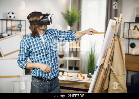 Vue latérale d'un artiste talentueux portant un casque VR peint avec palette et pinceau sur chevalet en studio.Concept de personnes, de technologie et d'espace artistique. Banque D'Images