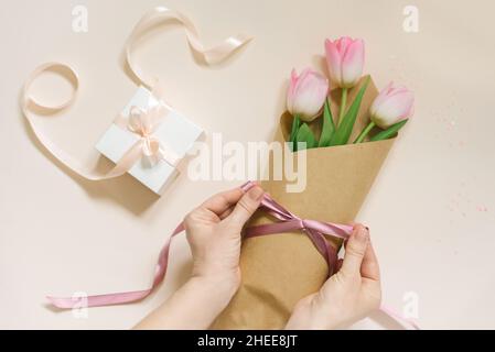 Les mains de femmes plates nouent un noeud en ruban satiné sur un simple bouquet de fleurs de tulipe rose frais en papier artisanal sur une table beige. Banque D'Images