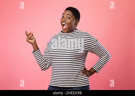 Une femme afro-américaine joyeuse de plus grande taille se croquant les doigts sur fond rose Banque D'Images