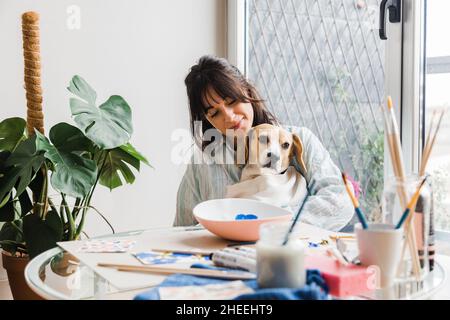 Belle femme peintre caressant mignon Beagle chien assis à la table avec un bol peint et divers pinceaux dans un studio léger Banque D'Images