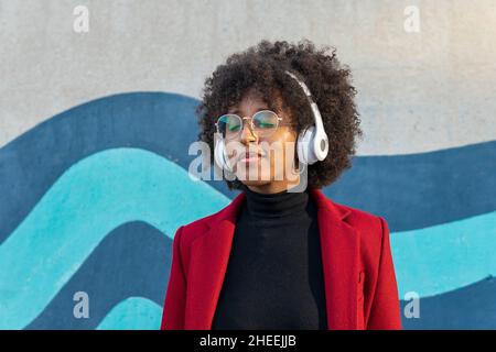 Femme afro-américaine confiante avec une coiffure afro et des lunettes portant un col de cygne noir et une veste rouge écoutant un podcast dans un casque sans fil Banque D'Images
