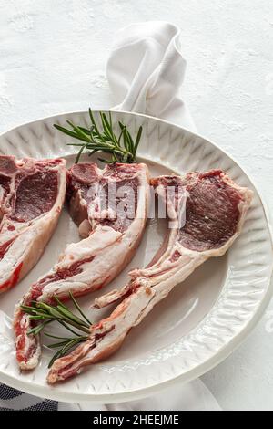 De dessus des côtelettes d'agneau non cuites servies sur une plaque en céramique blanche avec du romarin vert frais sur la table dans la cuisine légère Banque D'Images