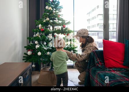 Une femme heureuse soldat en uniforme décorant l'arbre de Noël avec un fils sans visage dans un salon lumineux après son arrivée de la mission à la maison Banque D'Images