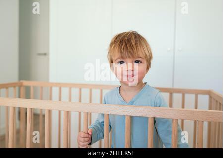 Adorable petit garçon aux cheveux blonds regardant l'appareil photo tout en se tenant dans un lit de bébé en bois dans une chambre lumineuse avec une garde-robe blanche à la maison Banque D'Images