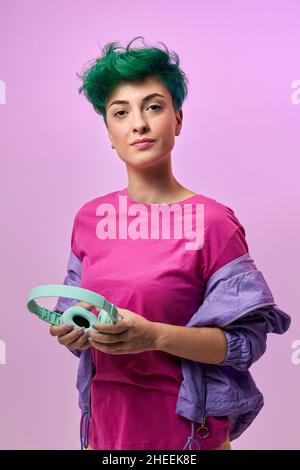 Positive millénaire femme avec casque dans la couleur rétro 80s tenue avec leurs mains tenant la veste et regardant loin sur violet Banque D'Images