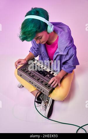 Corps complet d'une femme talentueuse en style rétro 80s et écouteurs jouant au clavier avec manette tout en composant de la musique sur le violet Banque D'Images