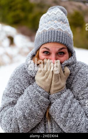 Une femme excitée portant un chapeau et des gants tricotés et une veste chaude qui gèle en hiver et qui couvre la bouche et le nez avec les mains Banque D'Images