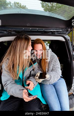 Des amies positives dans des vêtements chauds, assises sur le coffre ouvert de la voiture garée et défilant sur les médias sociaux sur le smartphone et parlant en hiver Banque D'Images