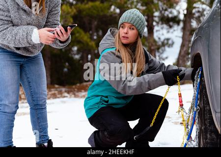 Des amies en casque chaud regardant les instructions sur le smartphone et le guide manuel essayant de mettre la chaîne de sécurité à neige sur le pneu de roue en squat Banque D'Images