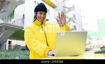 Homme asiatique souriant en vêtement d'extérieur et chapeau ayant la vidéoconférence sur netbook tout en se tenant dans la rue près du bâtiment résidentiel dans la ville Banque D'Images