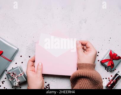 Les femmes tiennent la lettre d'amour vide message dans une enveloppe ouverte, carte de vœux de Saint-Valentin ou maquette vierge d'invitation.Espace de copie, vue de dessus.Fond rose avec coeurs, boîtes-cadeaux grises, confetti Banque D'Images