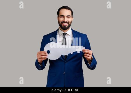 Homme barbu tenant à la main grande clé en papier regardant l'appareil photo avec un sourire crasseux, l'immobilier, son propre hébergement, portant un costume de style officiel.Prise de vue en studio isolée sur fond gris. Banque D'Images