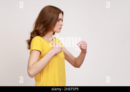 Portrait de profil d'une femme sérieuse préoccupée de jeune âge tenant des poings serrés prêts à la boxe, des cours d'auto-défense féminins, esprit de combat.Prise de vue en studio isolée sur fond gris. Banque D'Images