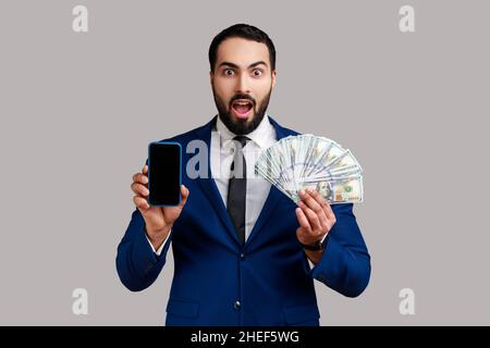 Un homme d'affaires barbu choqué qui détient des billets de banque en dollars et un téléphone portable avec écran vide maquette, portant un costume de style officiel.Prise de vue en studio isolée sur fond gris. Banque D'Images