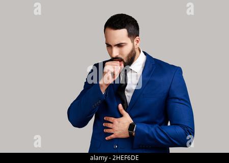 Portrait d'un homme barbu malsain toussant, attrape froid, ayant une température élevée, ayant un symptôme de grippe, portant un costume de style officiel.Prise de vue en studio isolée sur fond gris. Banque D'Images