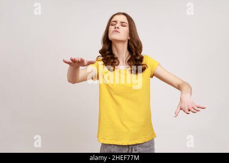 Portrait d'une adolescente aveugle à poil brun désorienté dans un T-shirt jaune décontracté marchant avec les yeux fermés, en tirant les mains pour trouver le chemin perdu.Prise de vue en studio isolée sur fond gris. Banque D'Images