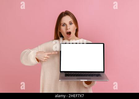 Portrait d'une femme blonde stupéfait regardant un appareil photo à bouche ouverte, pointant vers l'écran d'ordinateur portable avec écran blanc blanc, portant un chandail blanc.Studio d'intérieur isolé sur fond rose. Banque D'Images