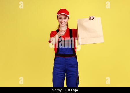 Portrait d'une femme de messagerie tenant le colis commandé, livrant la nourriture dans un sac en papier, pointant le doigt vers l'appareil photo, portant une combinaison et un chapeau.Studio d'intérieur isolé sur fond jaune. Banque D'Images