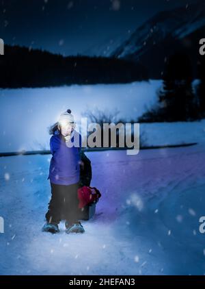 Canmore Alberta Canada, janvier 10 2021 : un enfant tirant un traîneau vers le haut d'une colline enneigée dans l'obscurité avec un phare à DEL pendant une tempête d'hiver. Banque D'Images