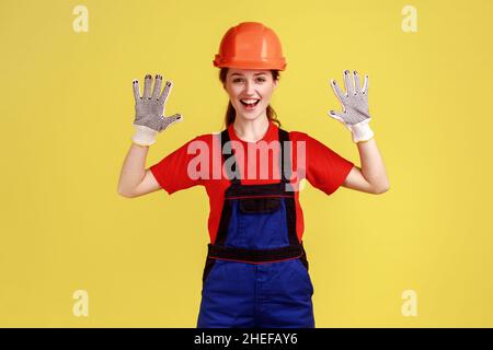 Portrait d'une travailleuse excitée debout avec des bras levés, montrant ses gants pour protéger les mains pendant le travail porte une combinaison et un casque de protection.Studio d'intérieur isolé sur fond jaune Banque D'Images