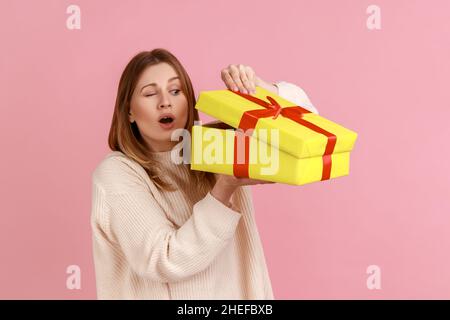 Portrait de jeune femme blonde curieuse attrayante regardant à l'intérieur jaune boîte de cadeau, étant intéressé ce à l'intérieur, portant chandail blanc.Studio d'intérieur isolé sur fond rose. Banque D'Images