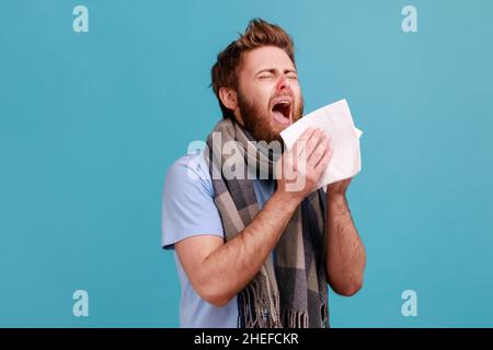 Épidémie de virus, épidémie de grippe.Homme enveloppé d'un foulard éternuant dans une serviette, nettoyant le nez et la toux, souffrant de symptômes grippaux, fièvre.Studio d'intérieur isolé sur fond bleu. Banque D'Images